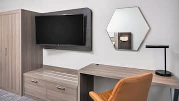 A modern hotel room with a flat-screen TV mounted on the wall, a wooden desk with drawers, a leather chair, and a hexagonal mirror.