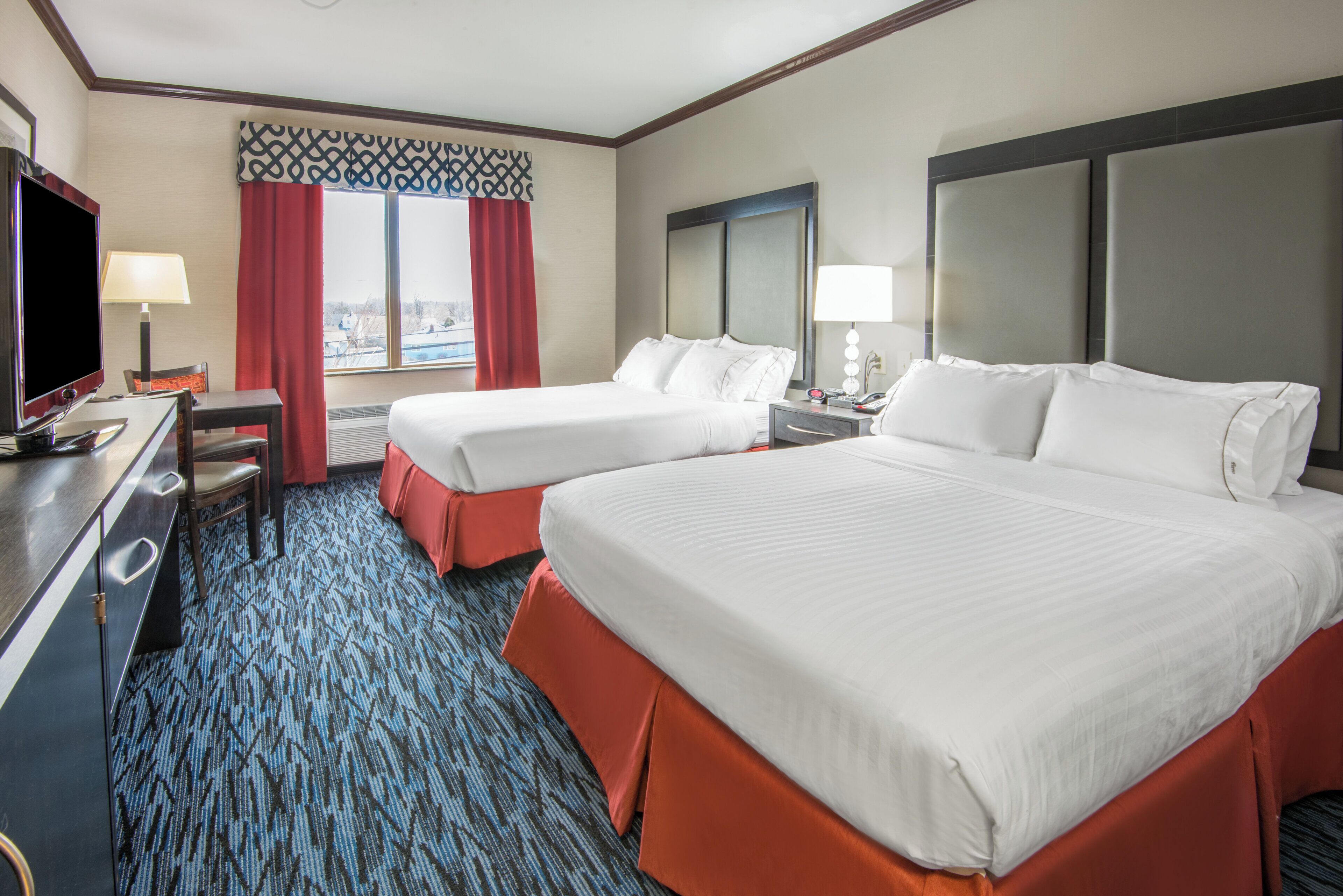 A hotel room with two beds, a desk with a television, and a window with red curtains.