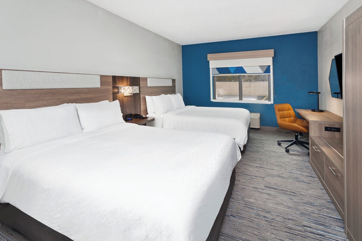 A hotel room with two beds, a desk, an orange chair, and a large window.