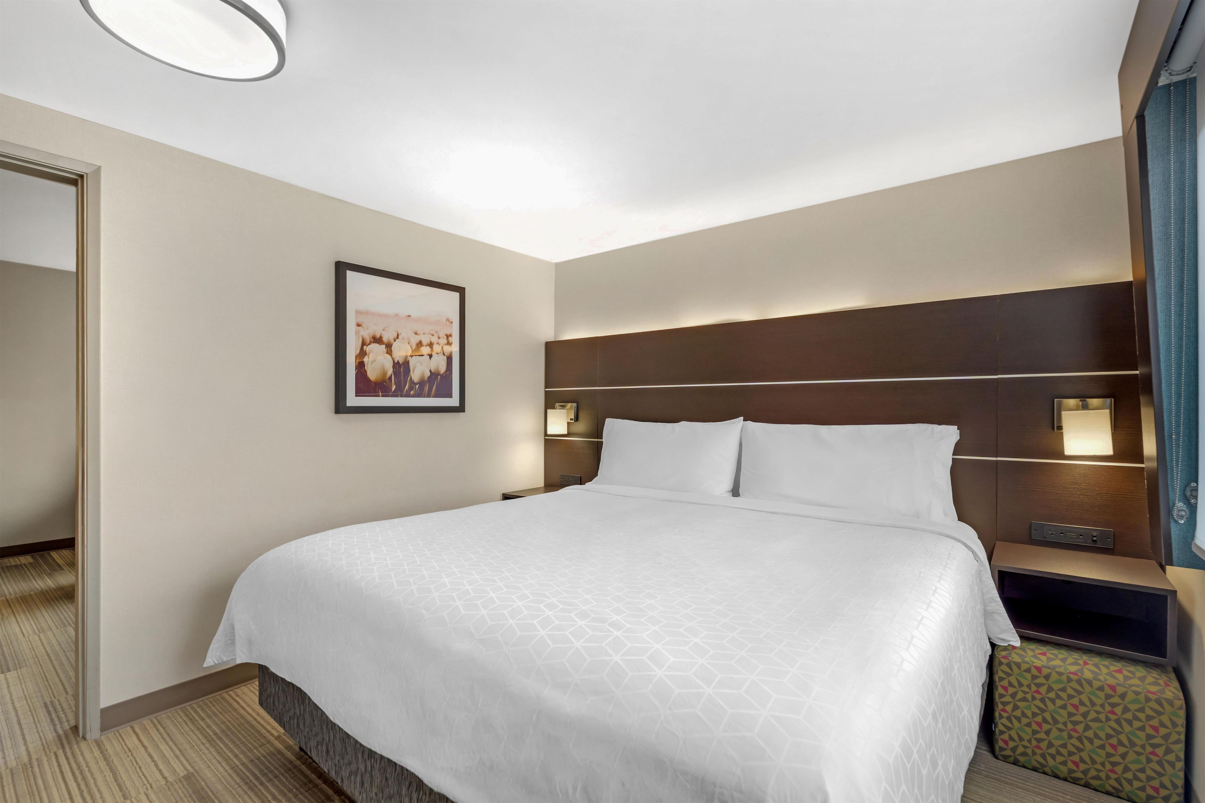 A hotel room with a large bed, a headboard with built-in lighting, a bedside table, and a framed picture on the wall.