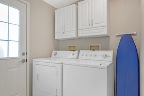 Une buanderie équipée de deux machines à laver à chargement frontal et d’un sèche-linge, d’un porte-serviettes avec une serviette bleue, et d’une fenêtre avec des stores.
