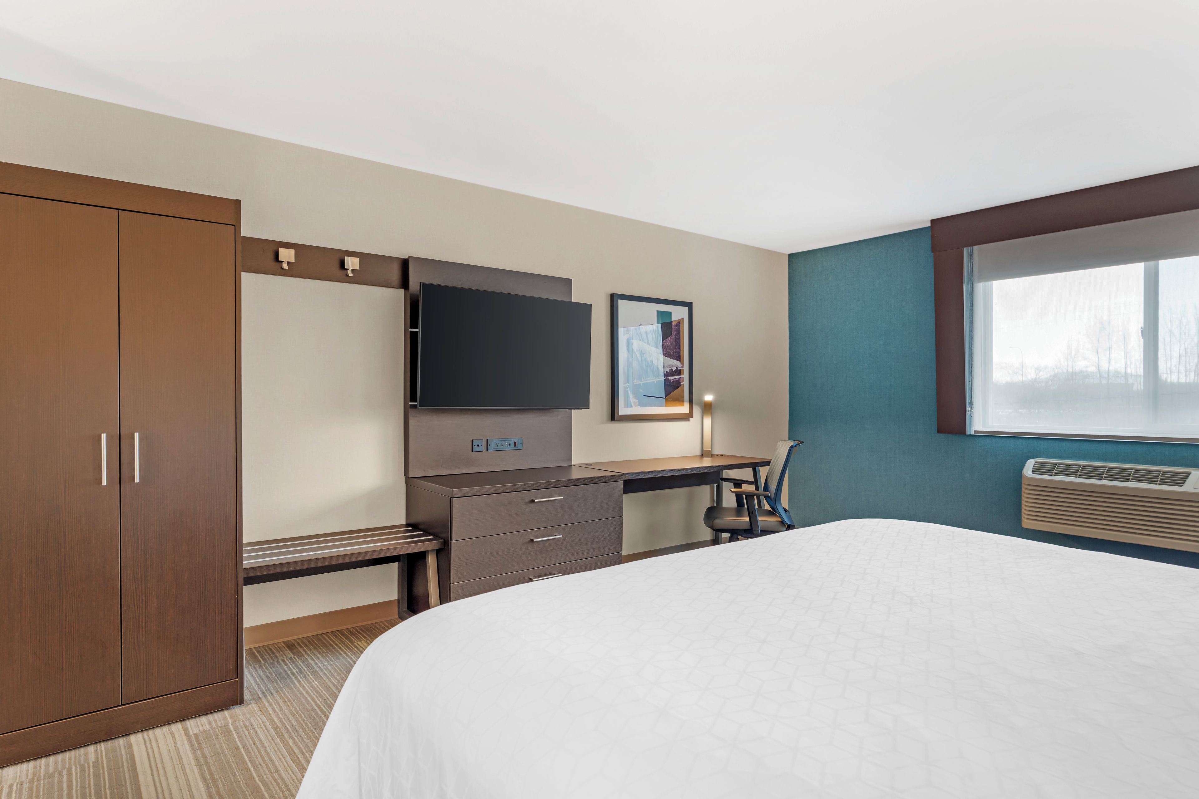 Ein Hotelzimmer mit Bett, Schreibtisch, Kleiderschrank, Flachbildfernseher und Klimaanlage.