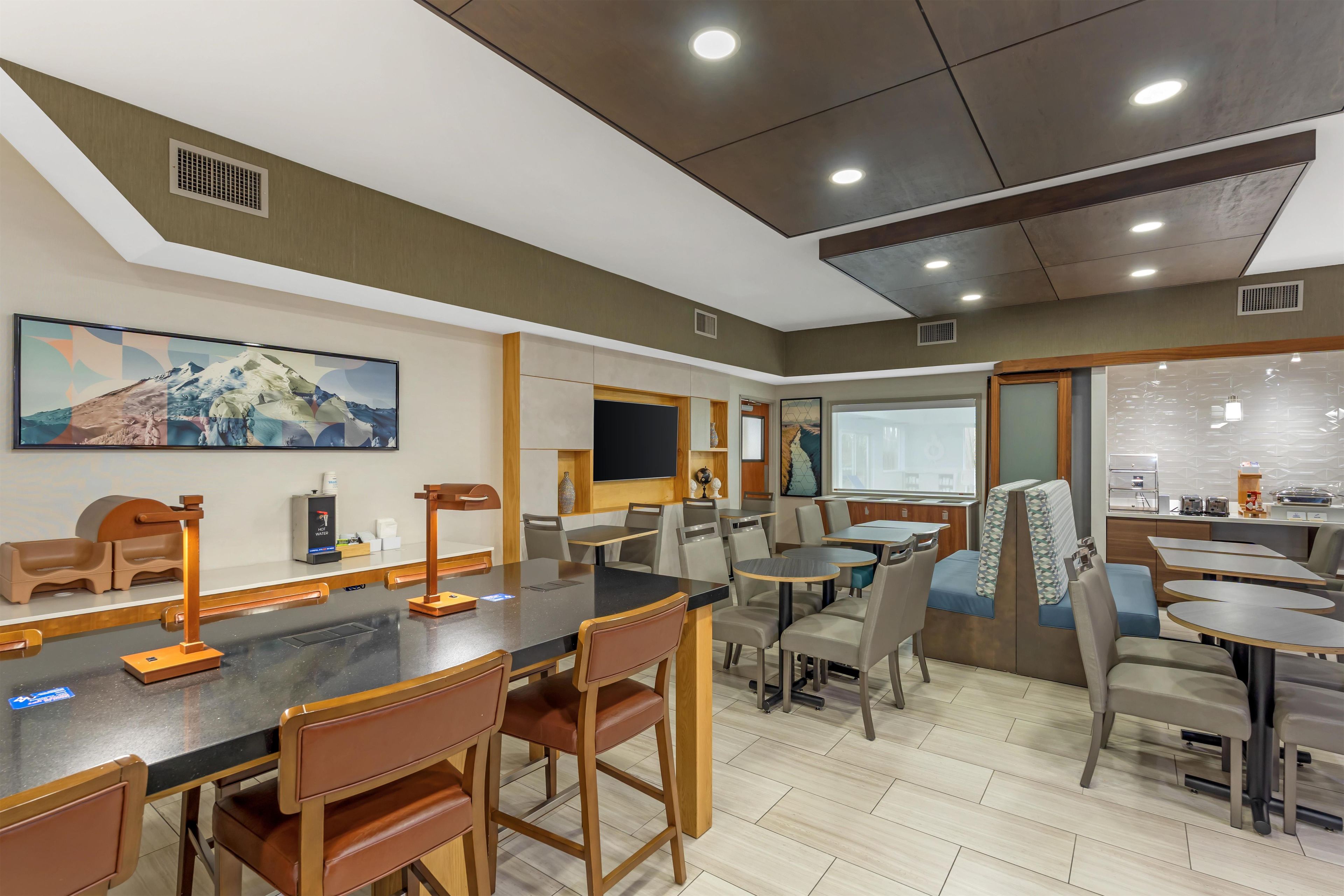 A modern dining area with multiple tables and chairs, a large painting on the wall, and a kitchen area visible through a glass partition.