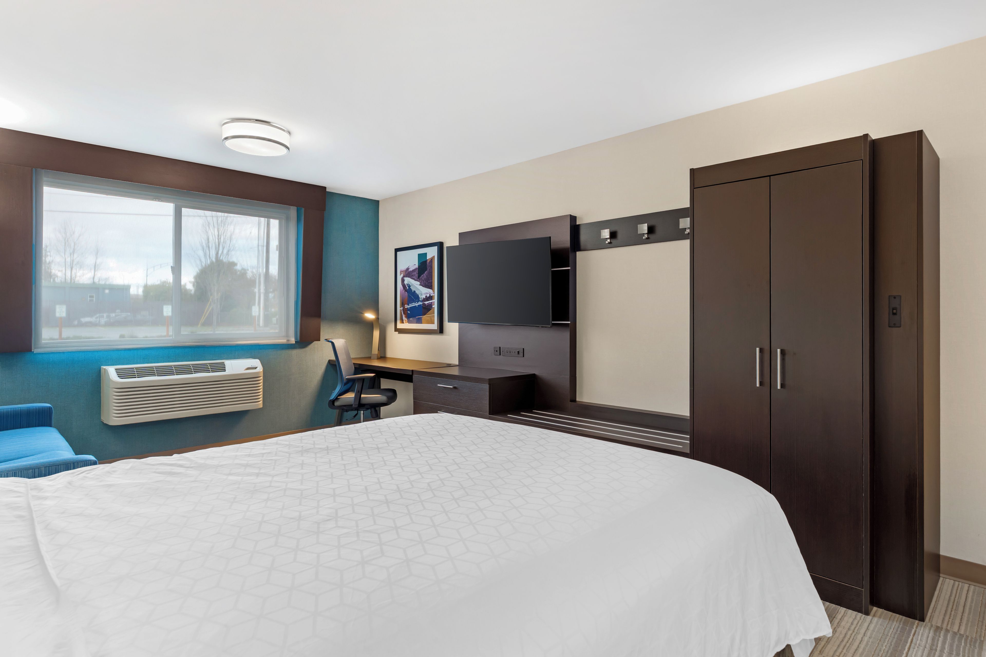 Une chambre d’hôtel comprenant un lit, un bureau avec une chaise, une télévision, une armoire et une fenêtre donnant sur l’extérieur.