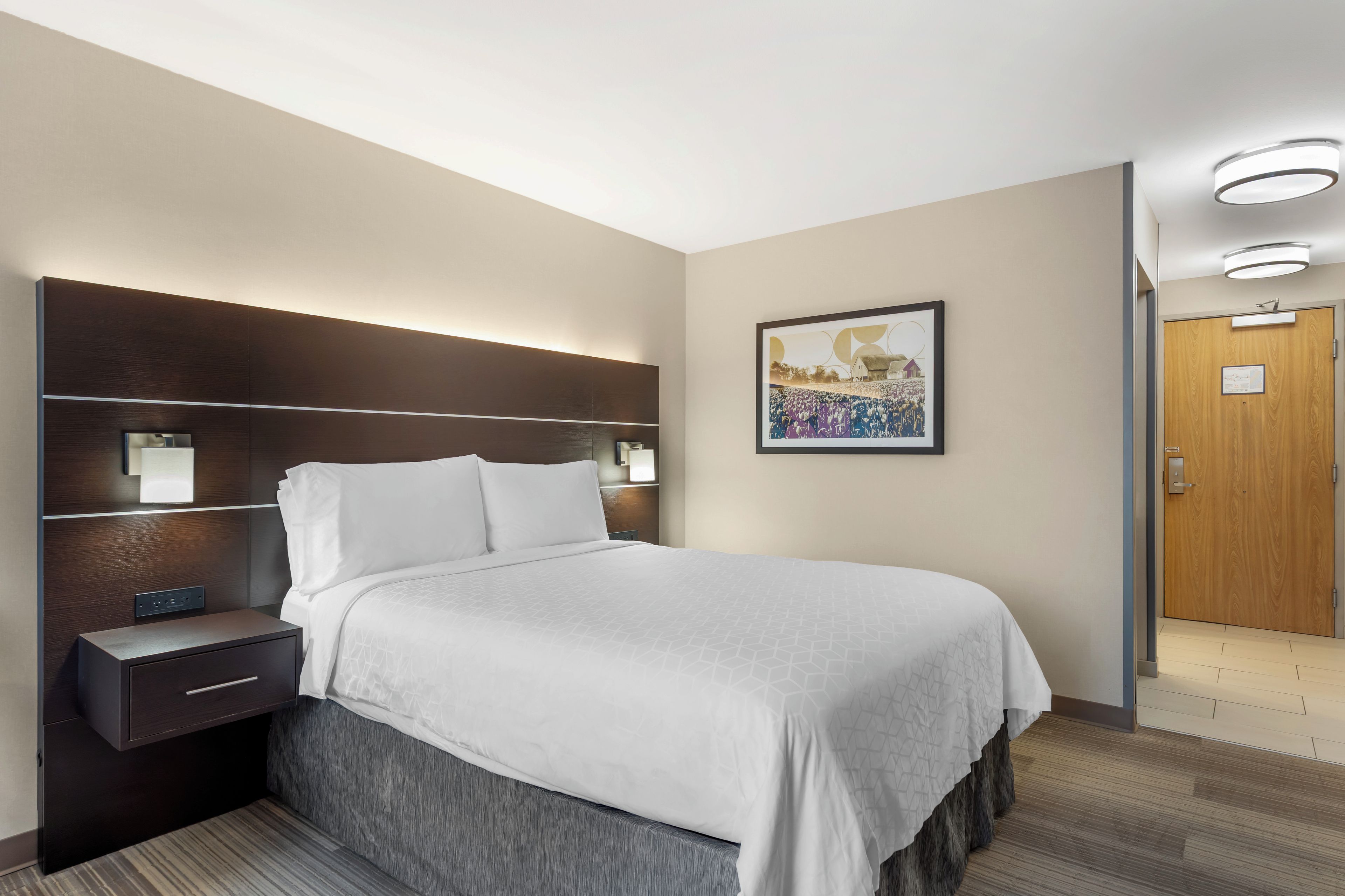 Une chambre d’hôtel comprenant un lit, des tables de chevet, une tête de lit, une œuvre d’art accrochée au mur et une porte.