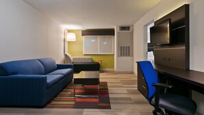 Ein modernes Hotelzimmer mit einem blauen Sofa, einem kleinen Tisch, einem Schreibtisch mit Stuhl, einem Fernseher und einem farbenfrohen, gestreiften Teppich.