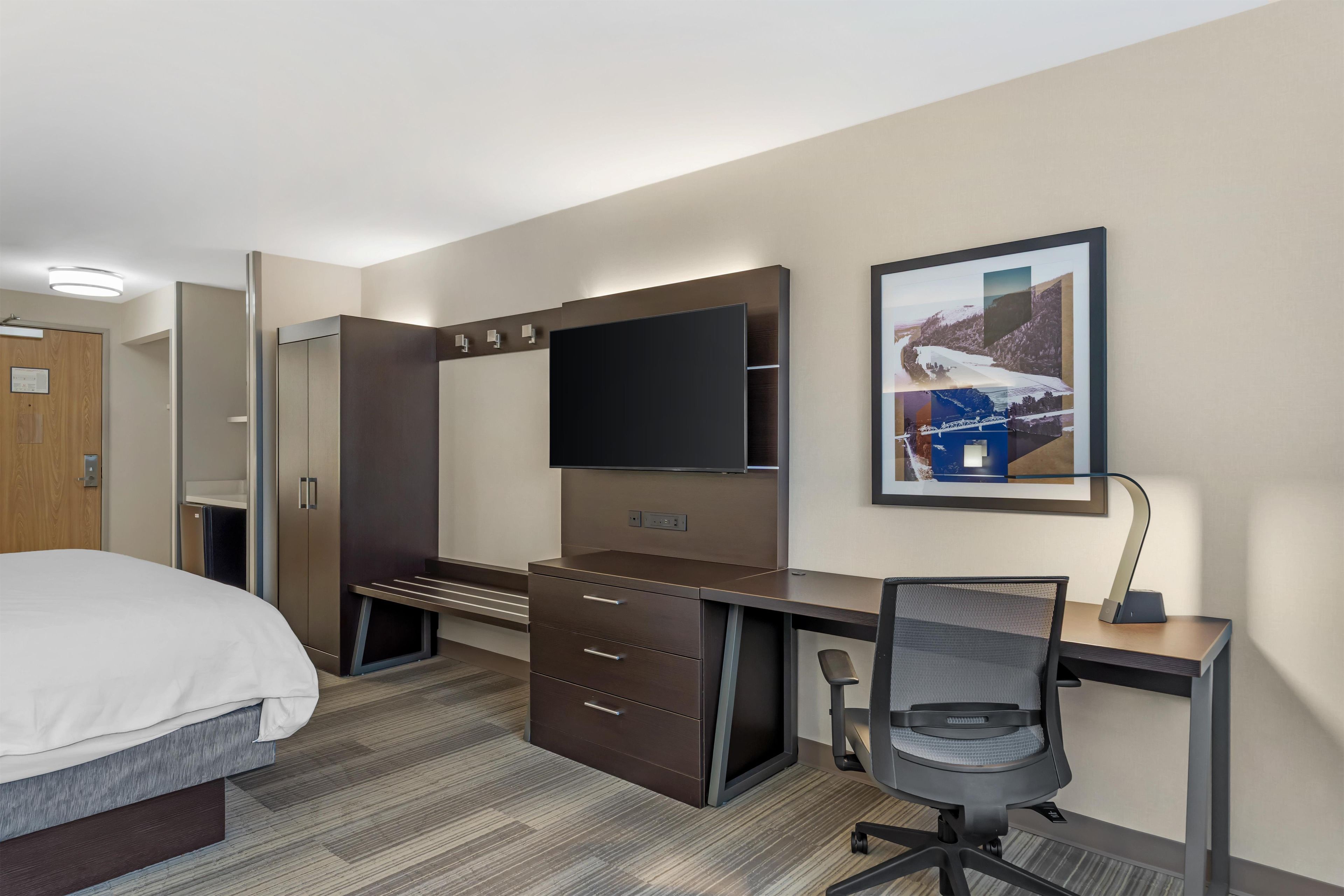 A hotel room with a bed, a desk with a chair, a TV, and a framed picture on the wall.