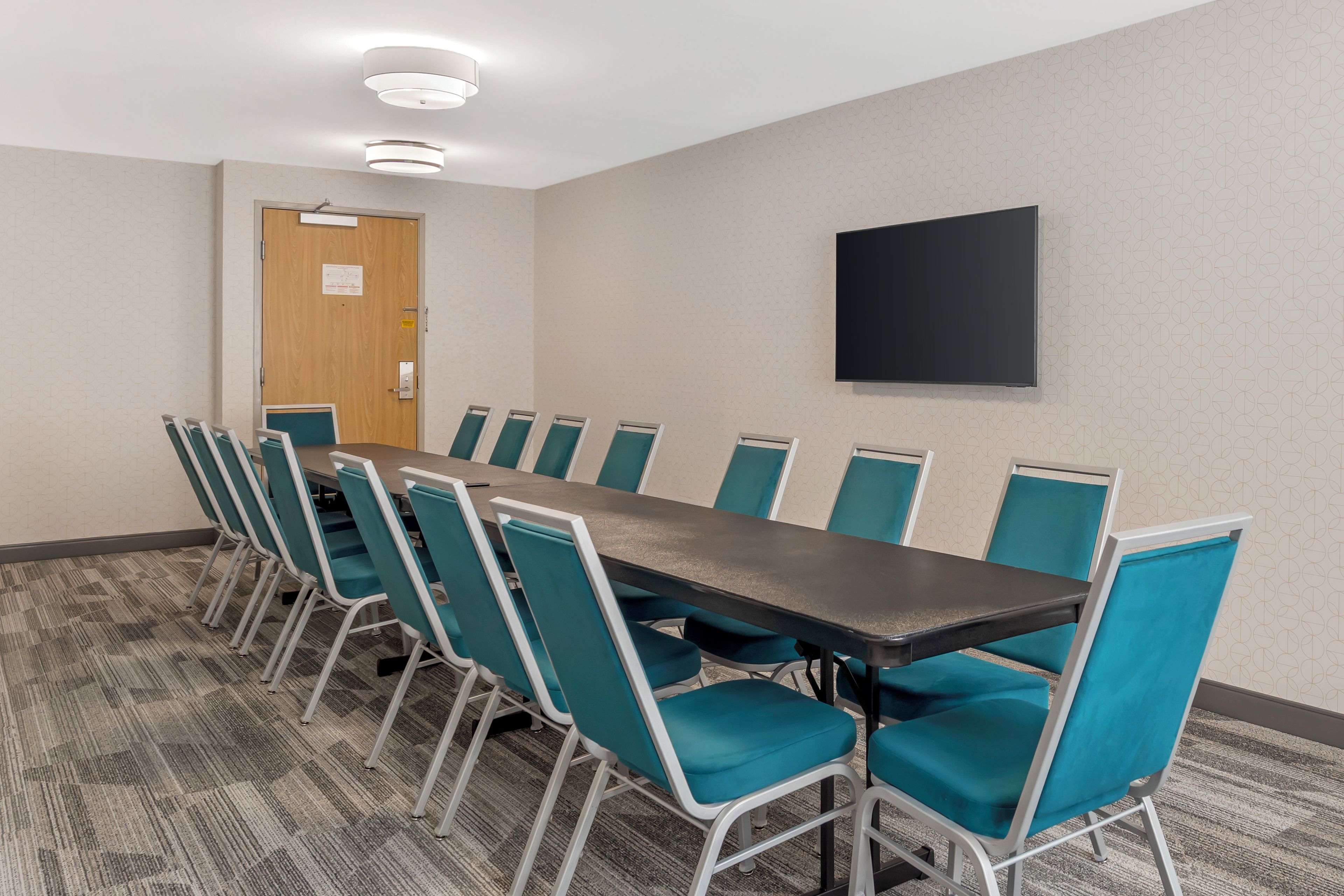 A conference room with a long table and chairs, a door, and a flat-screen TV on the wall.