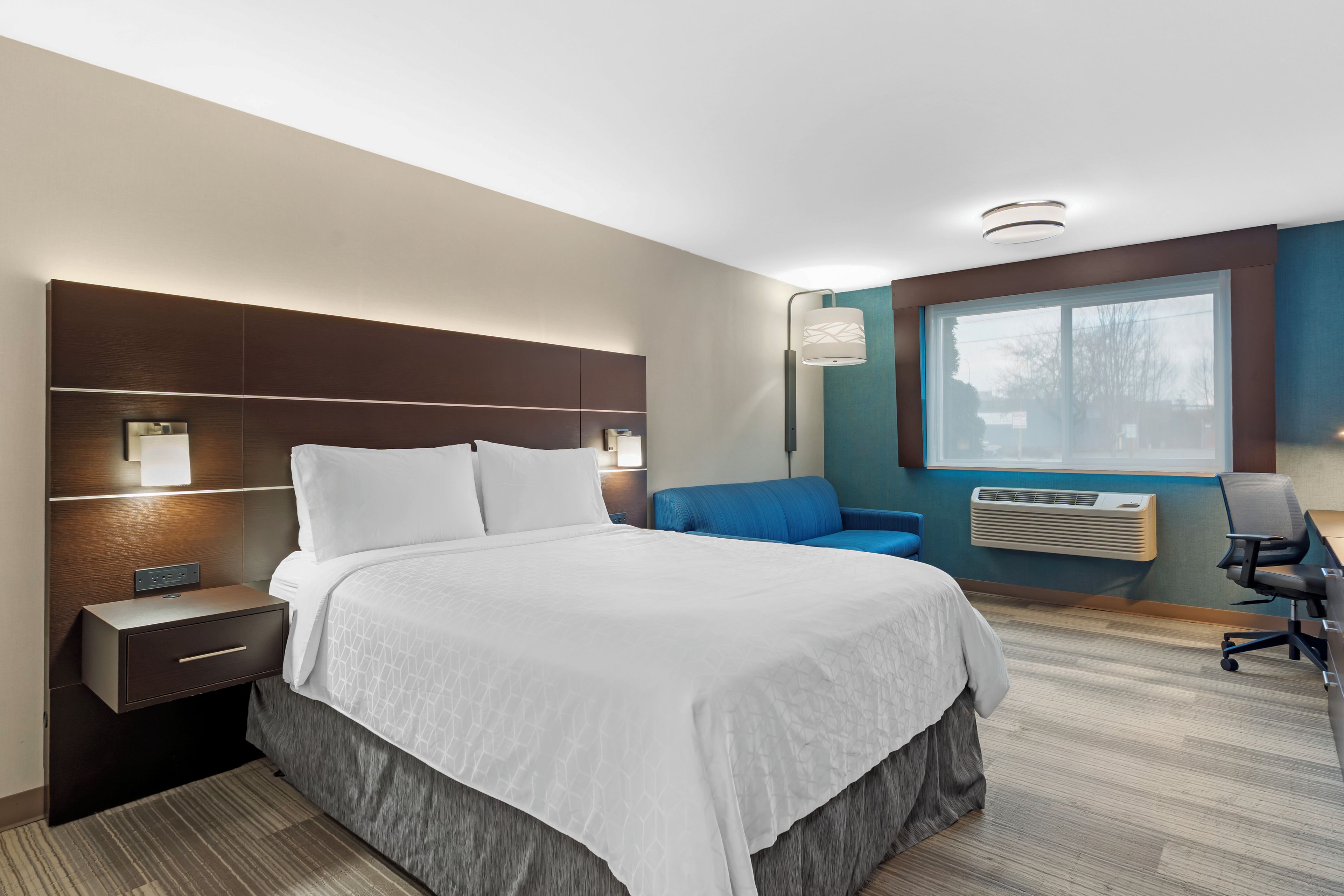 A hotel room with a large bed, bedside tables, a desk, a chair, a window, and an air conditioning unit.