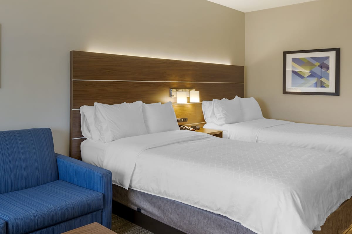 A hotel room with two beds, a blue chair, a wooden headboard, a nightstand with a lamp, and a framed abstract artwork on the wall.