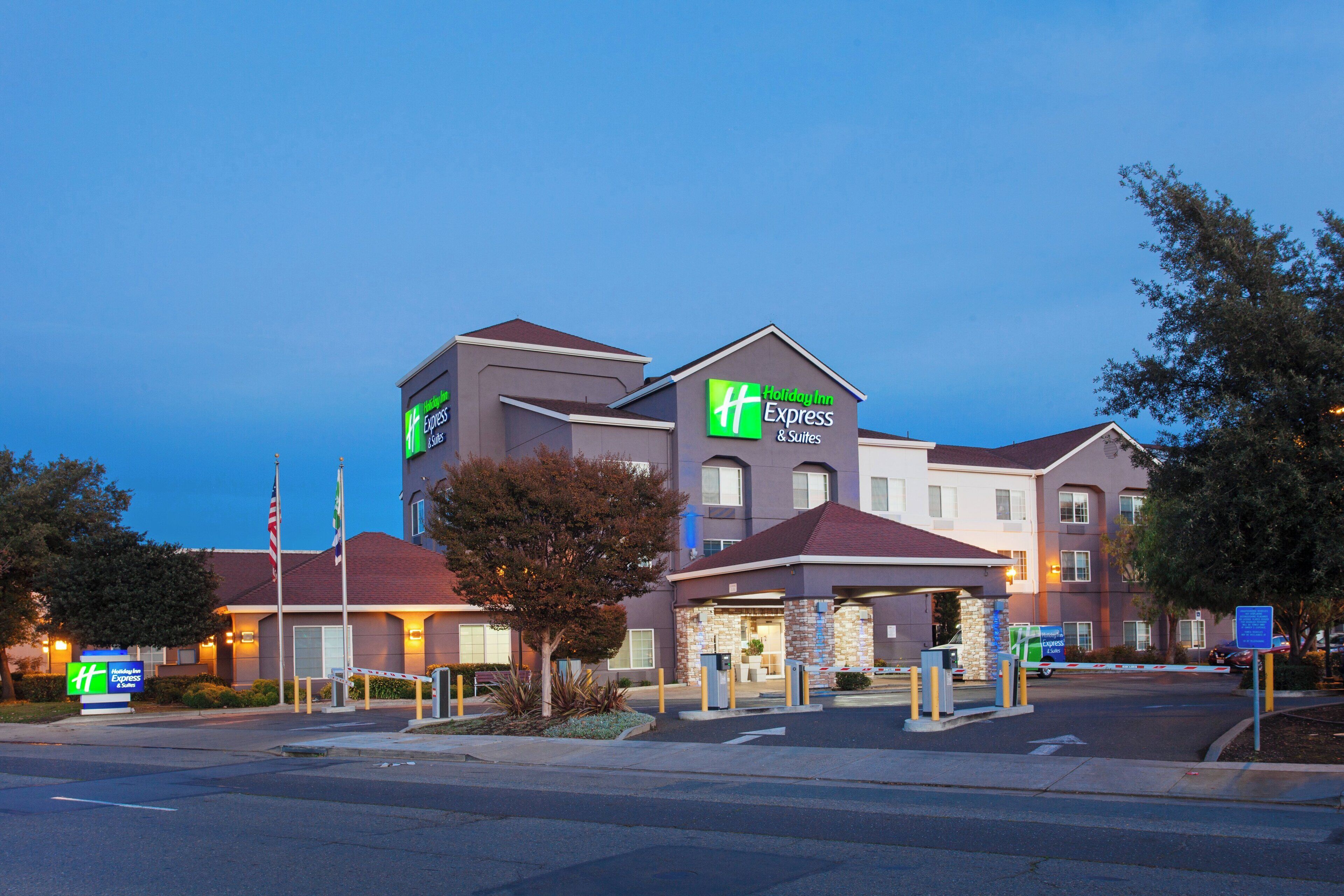 A Holiday Inn Express & Suites building with a modern design, featuring a prominent green and white logo, a parking lot, and a flagpole with the American flag.