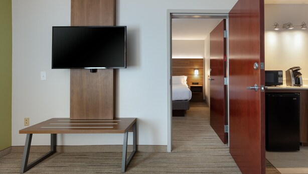 Een hotelkamer met een flatscreen televisie aan de muur, een houten bankje en een kitchenette met een magnetron en koffiezetapparaat.