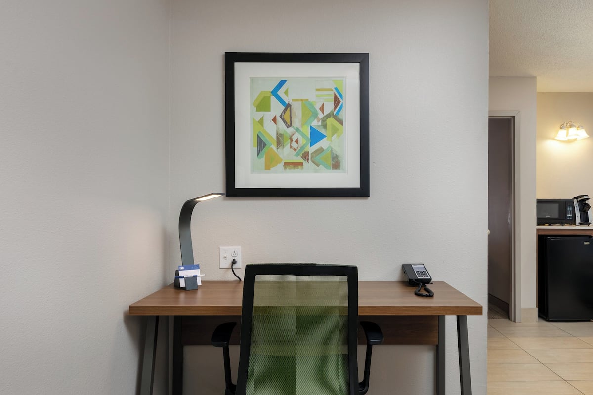 A hotel room with a wooden desk, a green mesh chair, a framed abstract painting, a desk lamp, and a telephone.