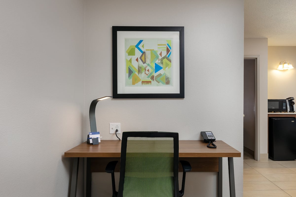 A hotel room with a wooden desk, a green mesh chair, a framed abstract painting, a desk lamp, and a telephone.