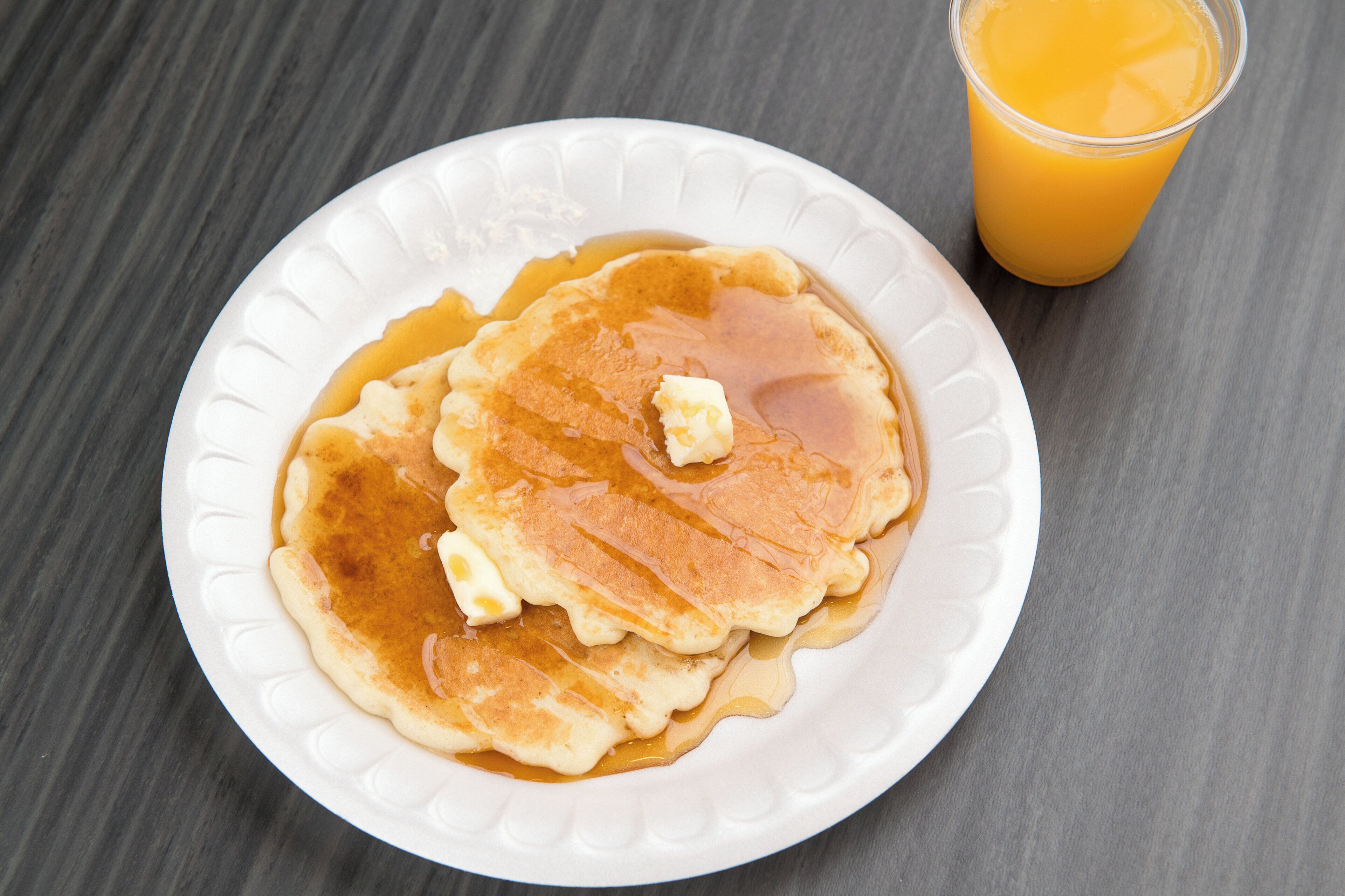 Une assiette de crêpes nappées de sirop et de beurre, posée sur une assiette blanche, avec un verre de jus d'orange en arrière-plan.
