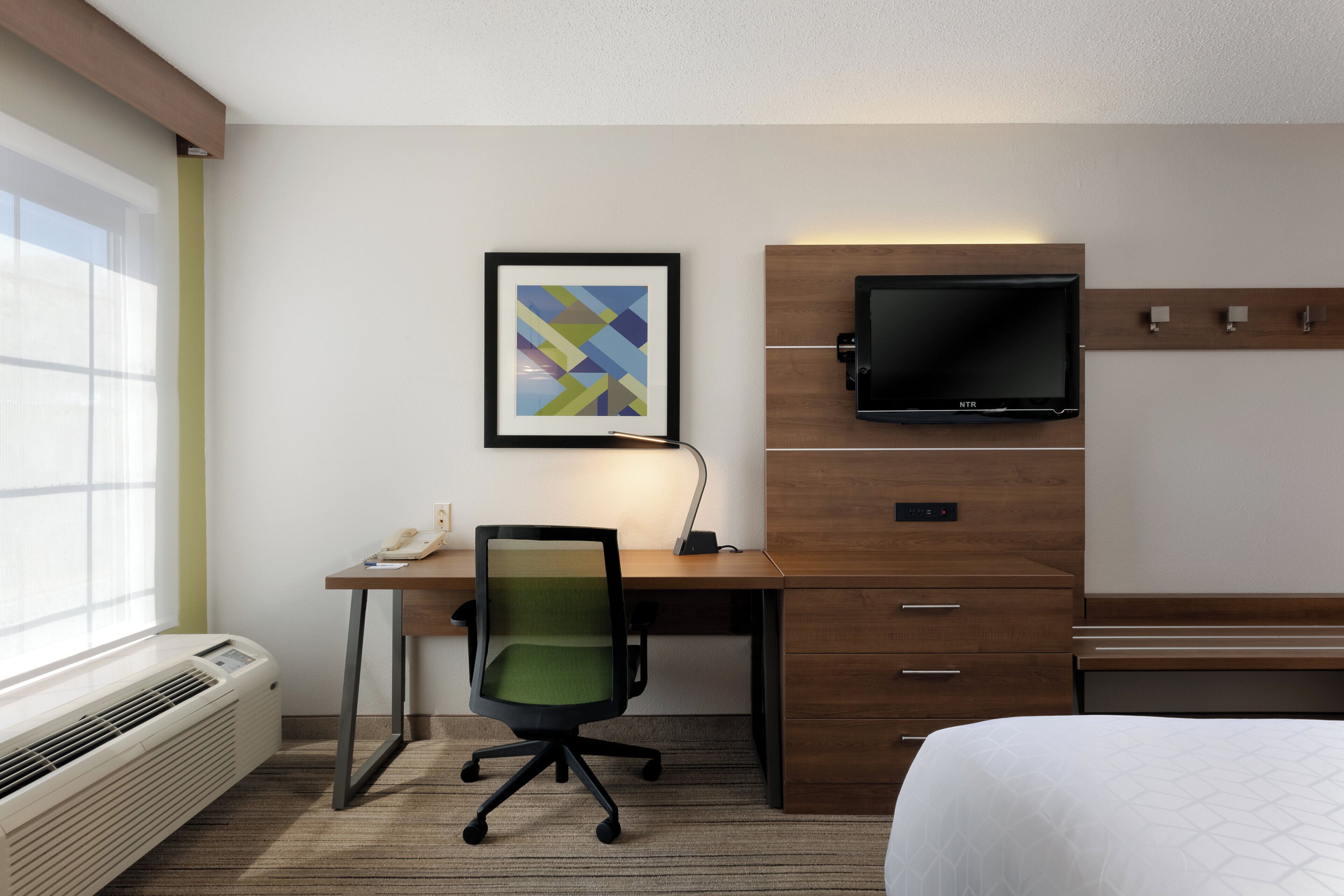 A hotel room with a desk, chair, bed, TV, and a framed picture on the wall.