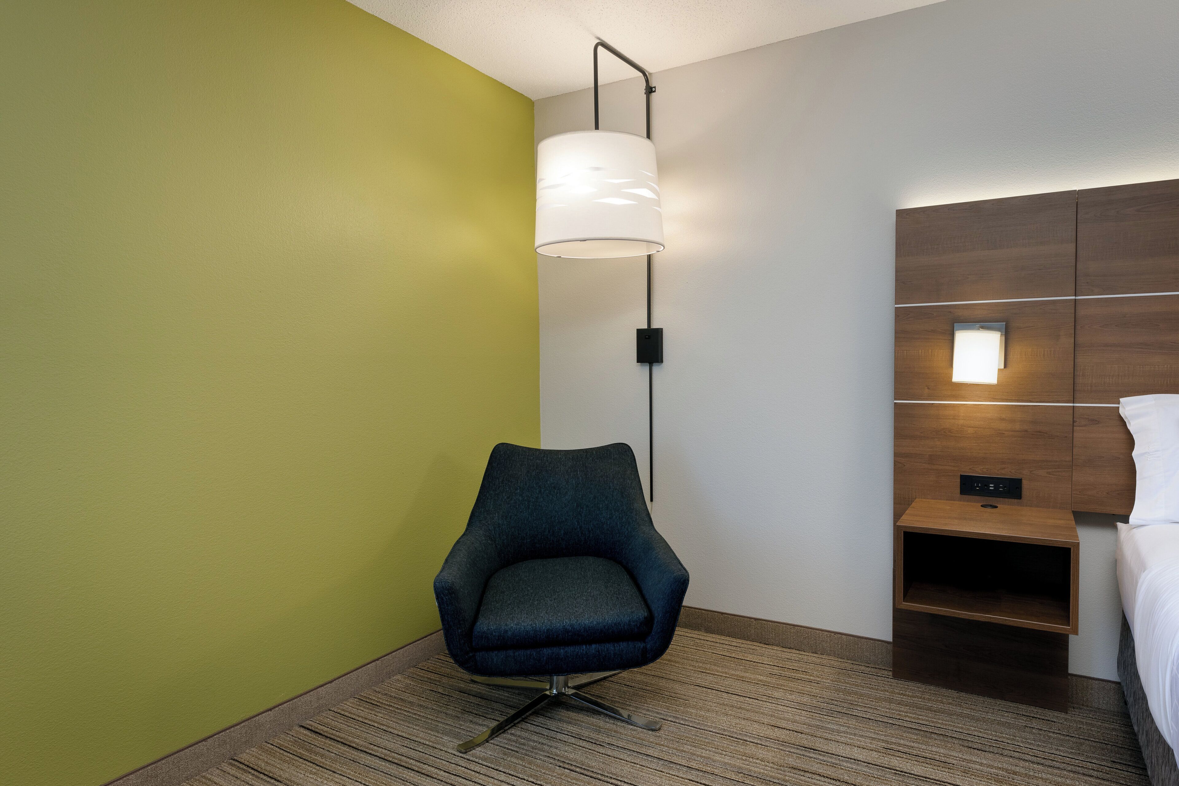Une chambre d’hôtel avec un mur vert, un fauteuil moderne, une table de chevet avec une lampe et une tête de lit en bois.