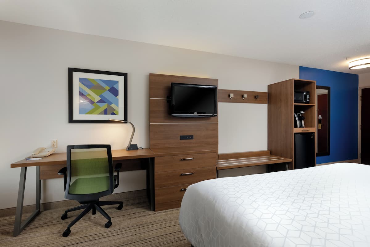 A hotel room with a wooden desk, a television mounted on the wall, a bed with a white bedspread, and a framed abstract artwork on the wall.