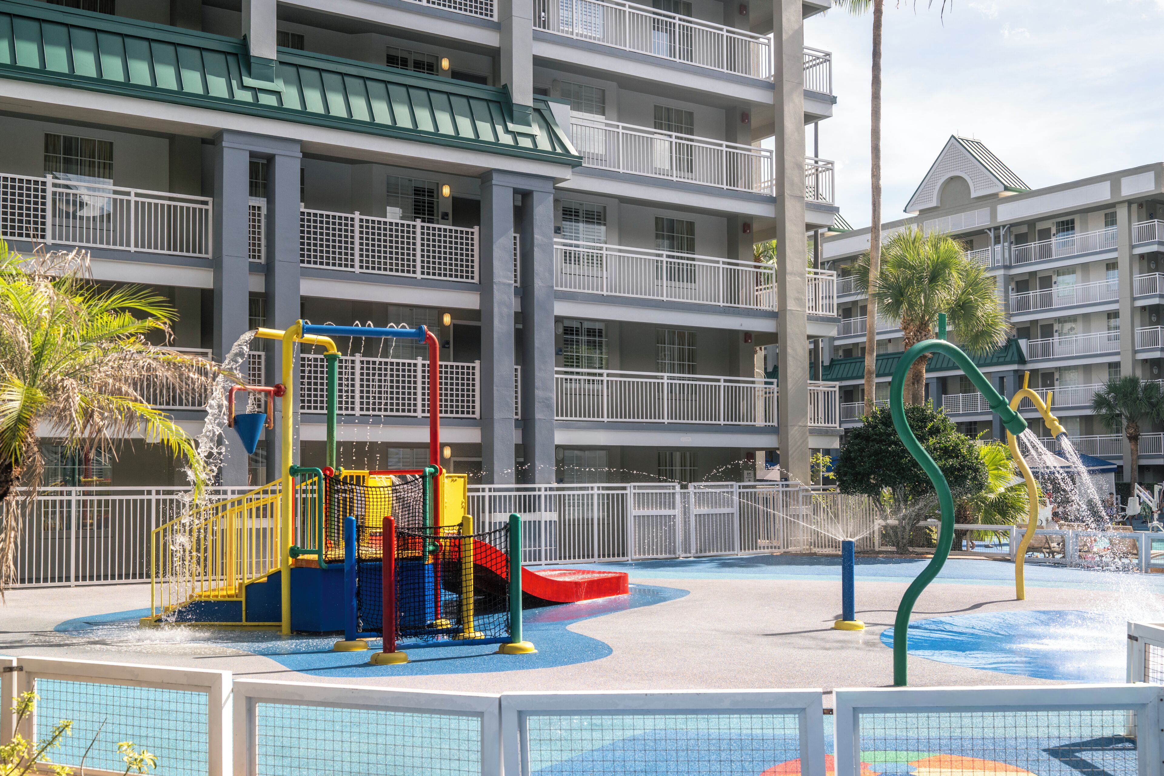 Un parque infantil con zona de juegos acuáticos, rodeado por un hotel de varios pisos con balcones y techo ajardinado.