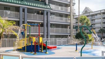 Um parquinho infantil com área de recreação aquática, rodeado por um hotel com várias andares, varandas e telhado verde.
