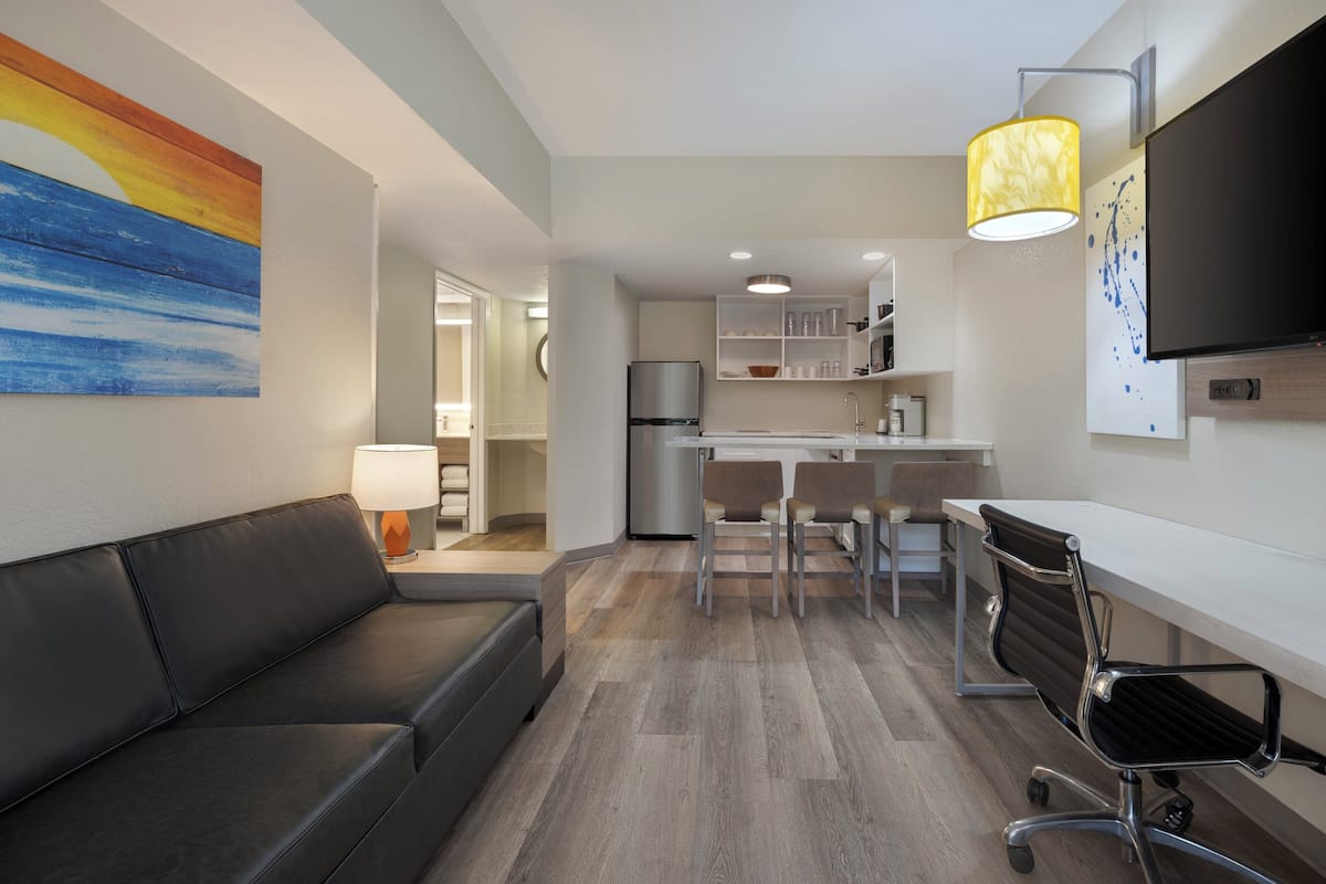 A modern living room with a black leather sofa, a dining area with a white table and chairs, a kitchen with white cabinets and a refrigerator, and a mounted flat-screen TV.