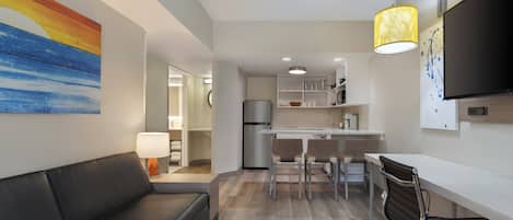 A modern living room with a black leather sofa, a dining area with a white table and chairs, a kitchen with white cabinets and a refrigerator, and a mounted flat-screen TV.