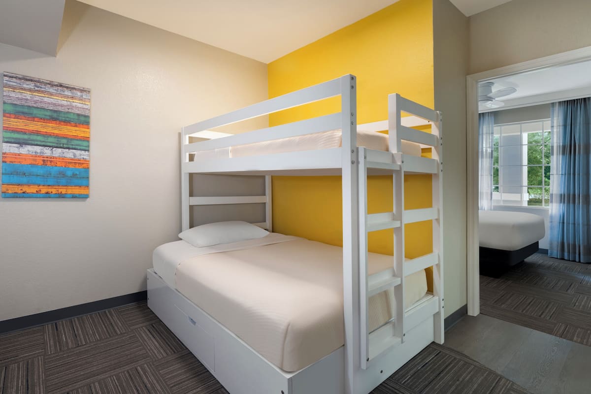 A bunk bed room with a window, a painting on the wall, and a bathroom visible through the window.
