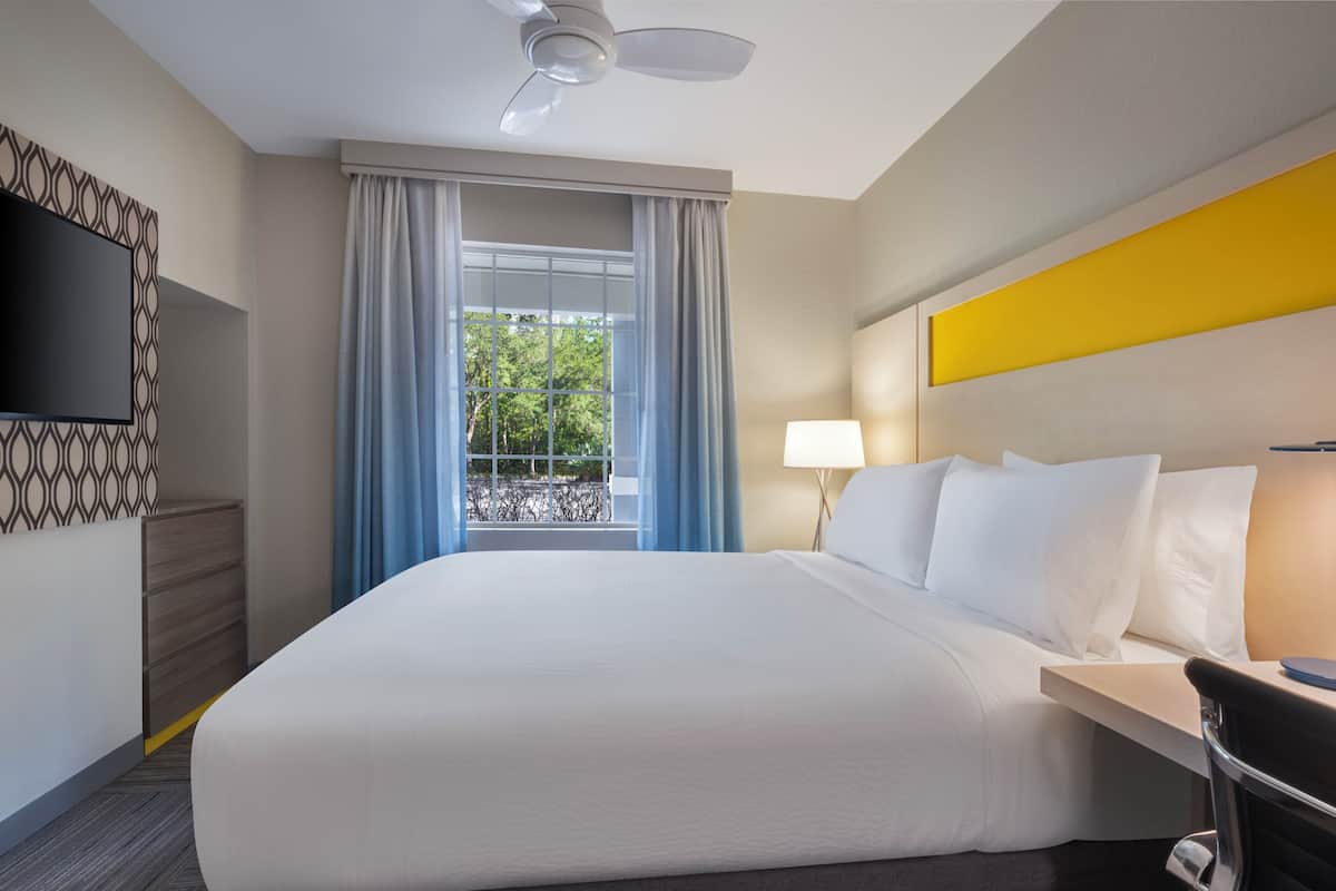 A hotel room with a bed, a TV, a window with curtains, a lamp, and a ceiling fan.