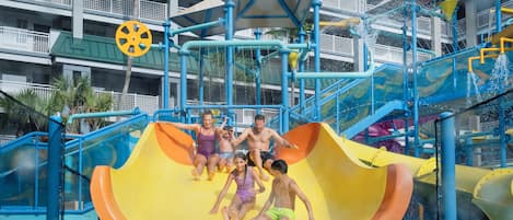Parque acuático con un tobogán amarillo, atracciones de agua en tonos azul y amarillo, y un edificio de varios niveles con balcones al fondo.