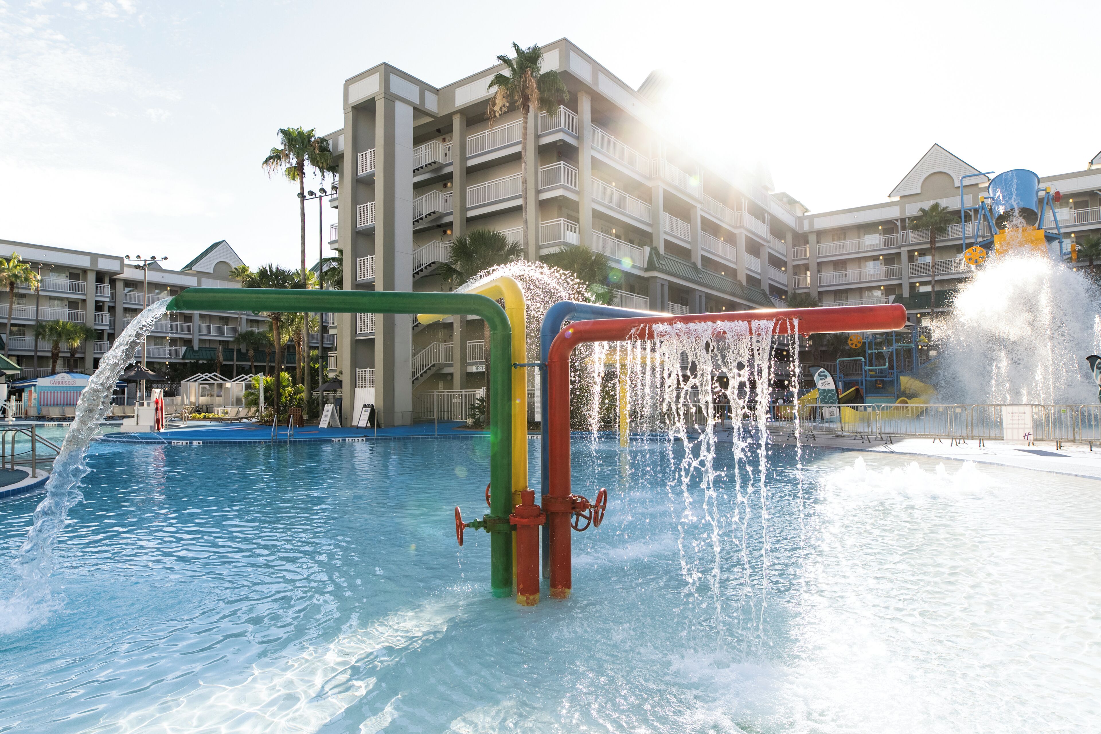 A water park with multiple water features, including a water slide and fountains, set against a backdrop of a hotel with balconies and palm trees.