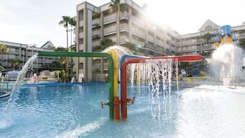Un parque acuático con diversas atracciones, incluyendo un tobogán de agua y fuentes, con un hotel y palmeras al fondo.
