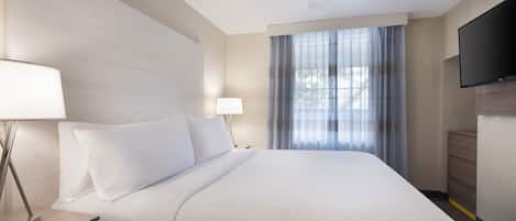 A hotel room with a bed, two bedside lamps, a ceiling fan, a TV, and a window with curtains.