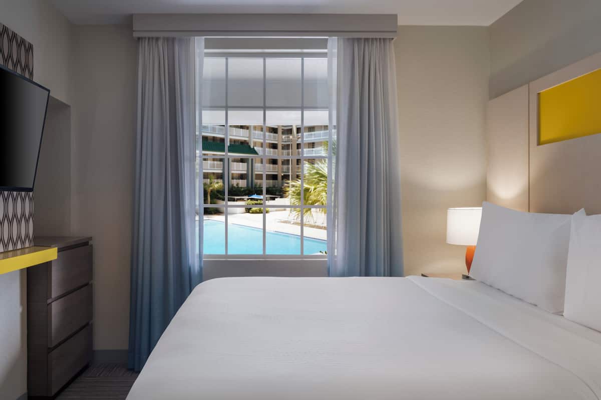 A hotel room with a bed, a TV, a desk, a lamp, and a window with a view of a pool and buildings.