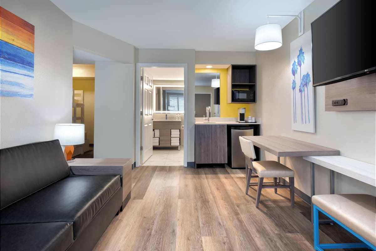 A modern hotel room with a sofa, a dining area, a kitchen, and a TV.