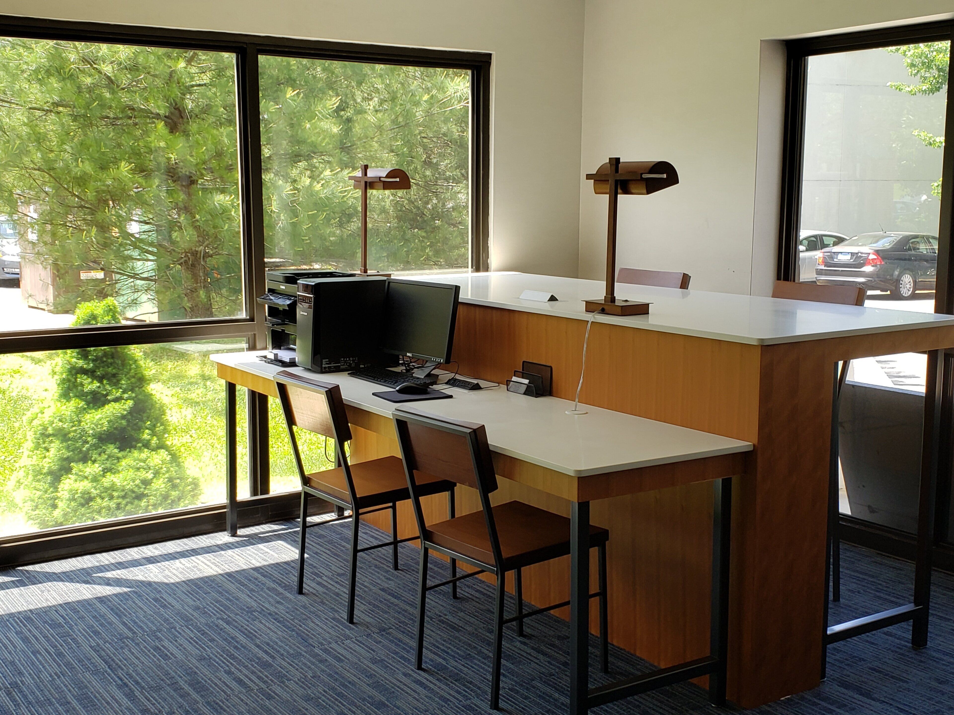A modern office space with a desk, computer, and two chairs. There are two standing lamps and a window with a view of trees and parked cars.