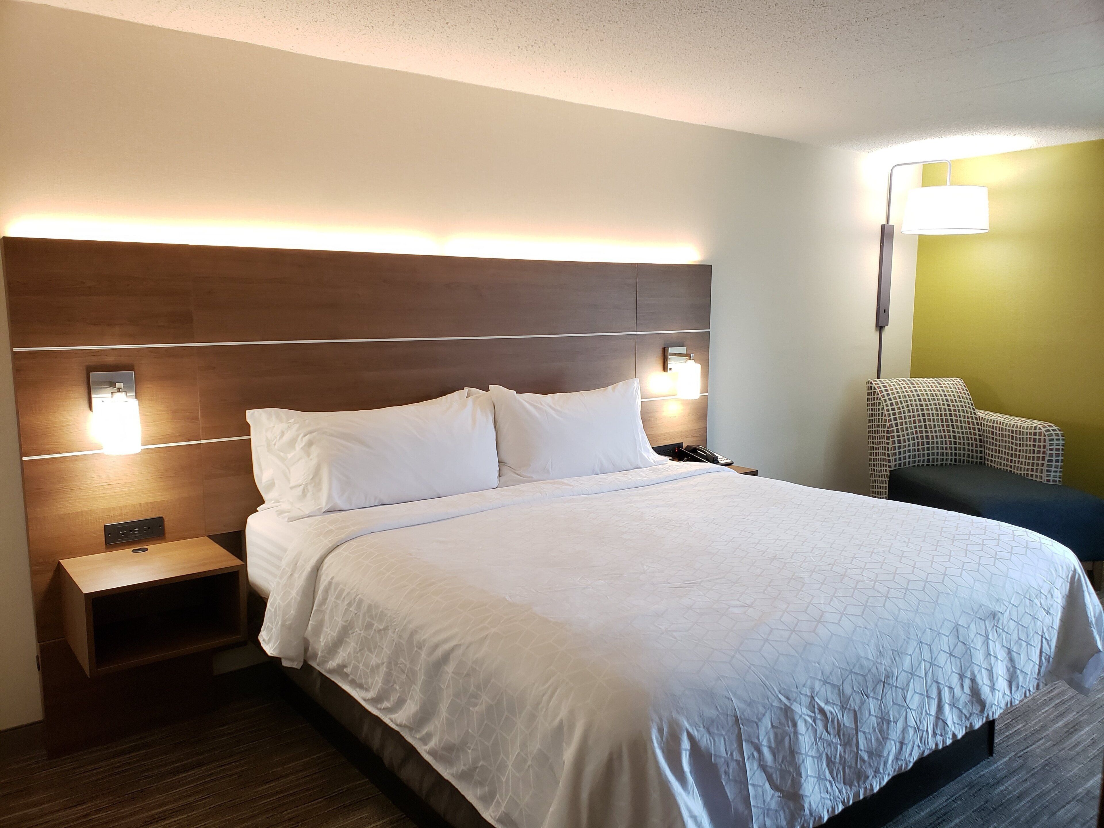 Una habitación de hotel con una cama grande, lámparas de noche, una mesita de noche, una silla y una lámpara de pared.