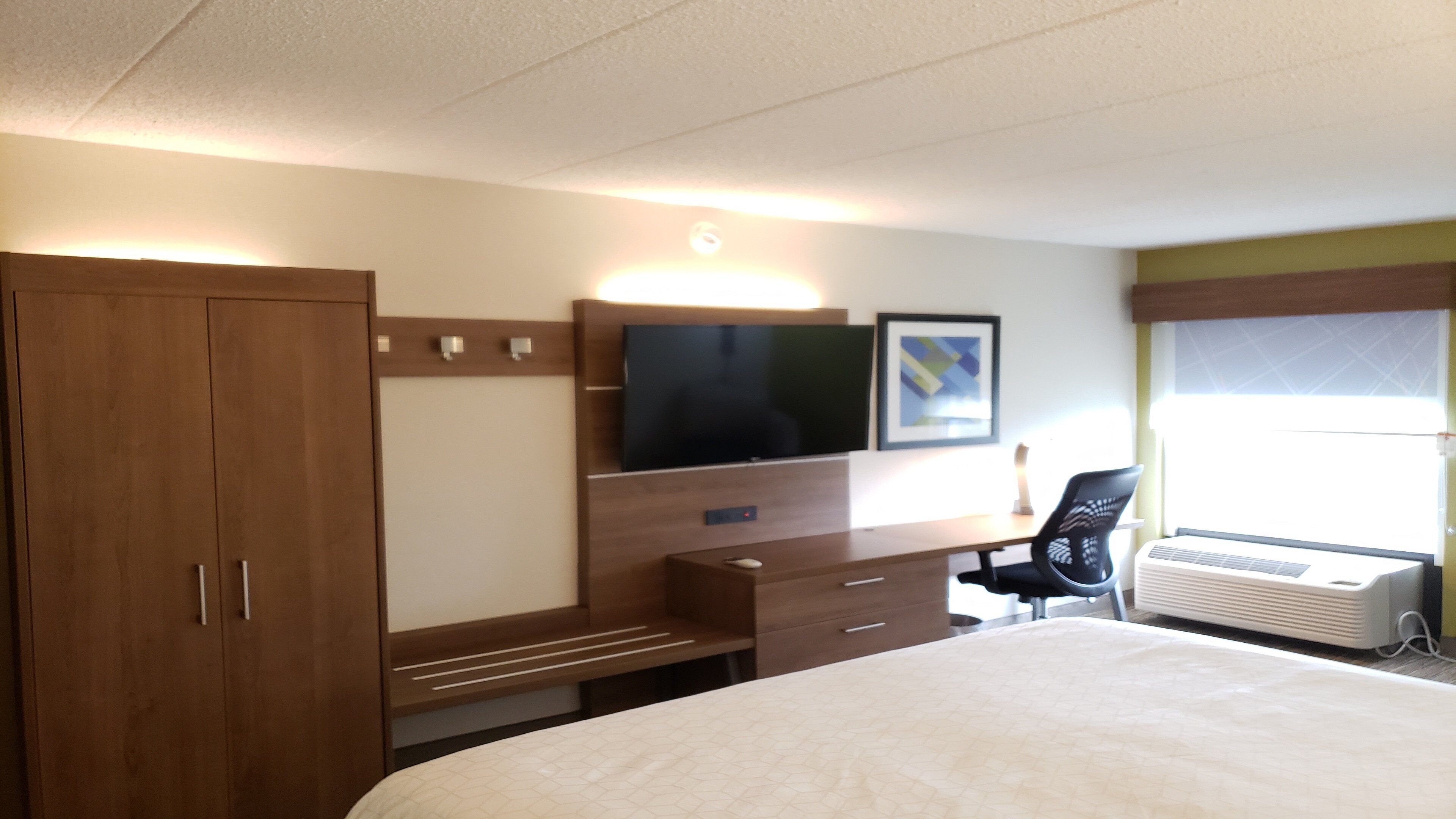 Una habitación de hotel con una cama, televisión montada en la pared, un escritorio con una silla y una ventana con persianas.