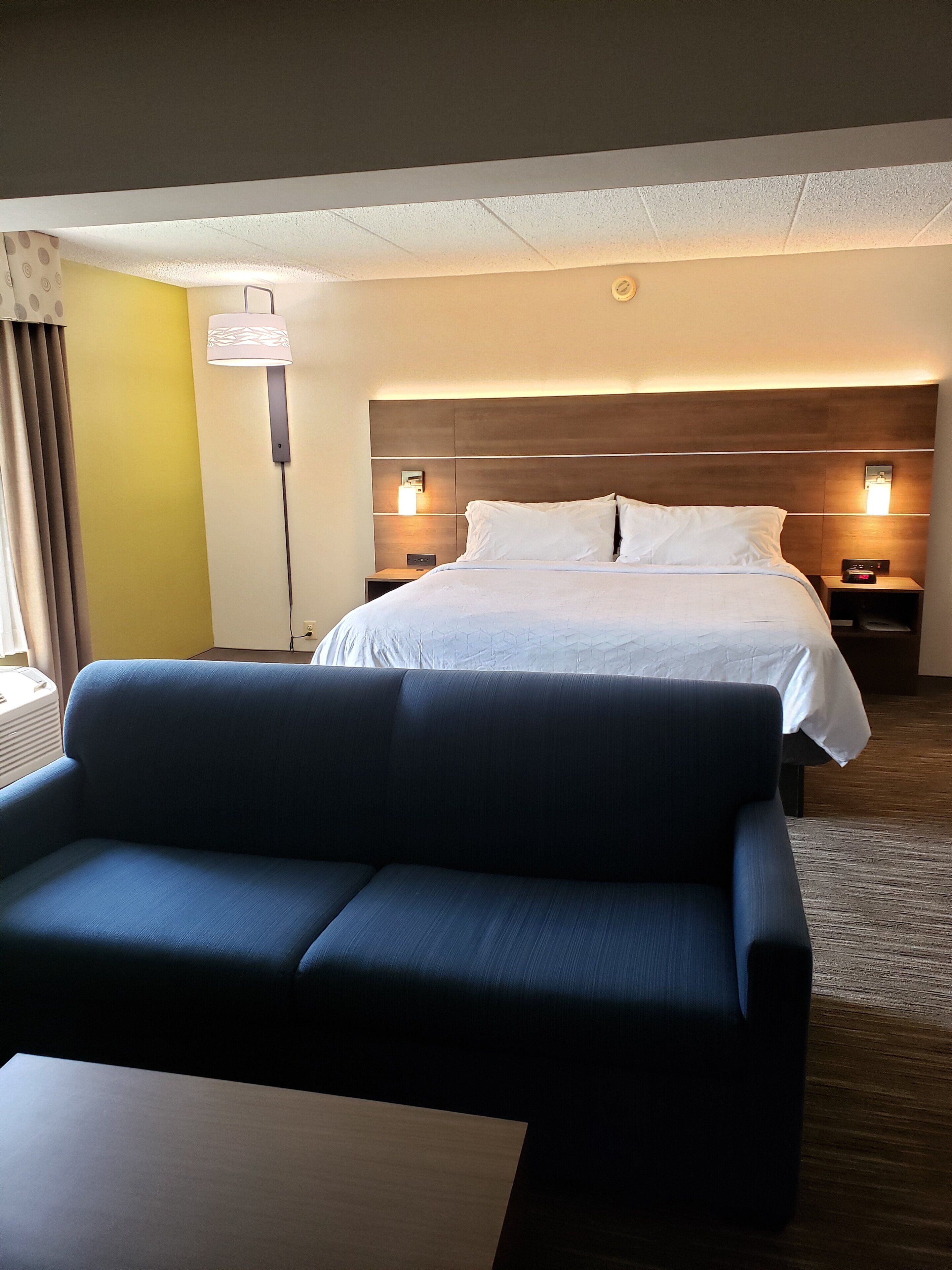 Habitación de hotel con cama, mesita de noche, lámpara, sofá y ventana con cortinas.