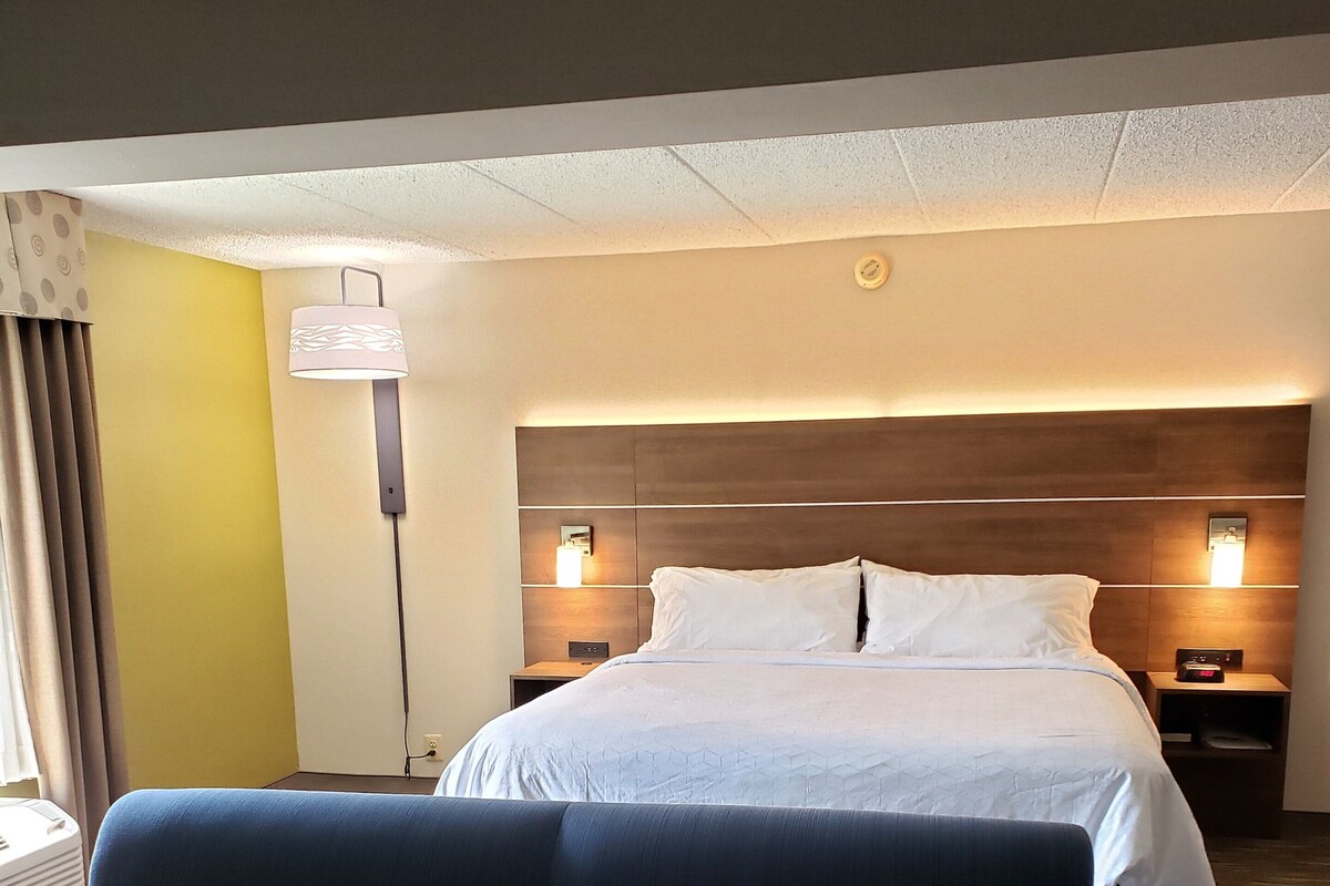 A hotel room with a bed, a nightstand, a lamp, a sofa, and a window with curtains.