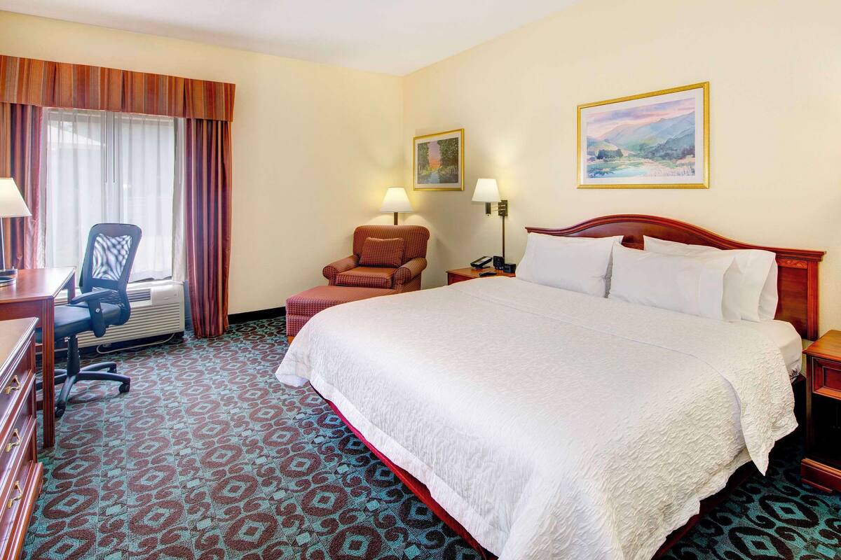 A hotel room with a large bed, a desk with a computer, a chair, a nightstand with a lamp, and two wall-mounted lamps.