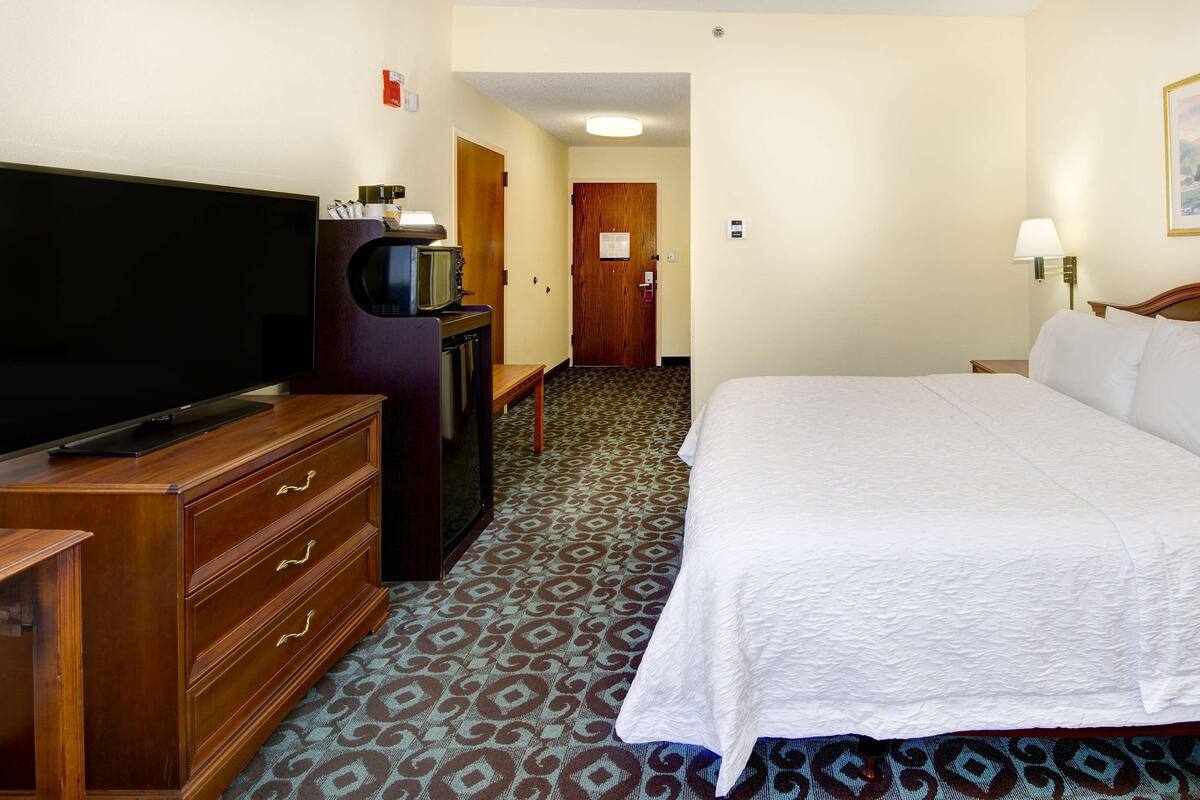 A hotel room with a large flat-screen TV, a wooden dresser, a bed with white linens, a patterned carpet, and a painting on the wall.