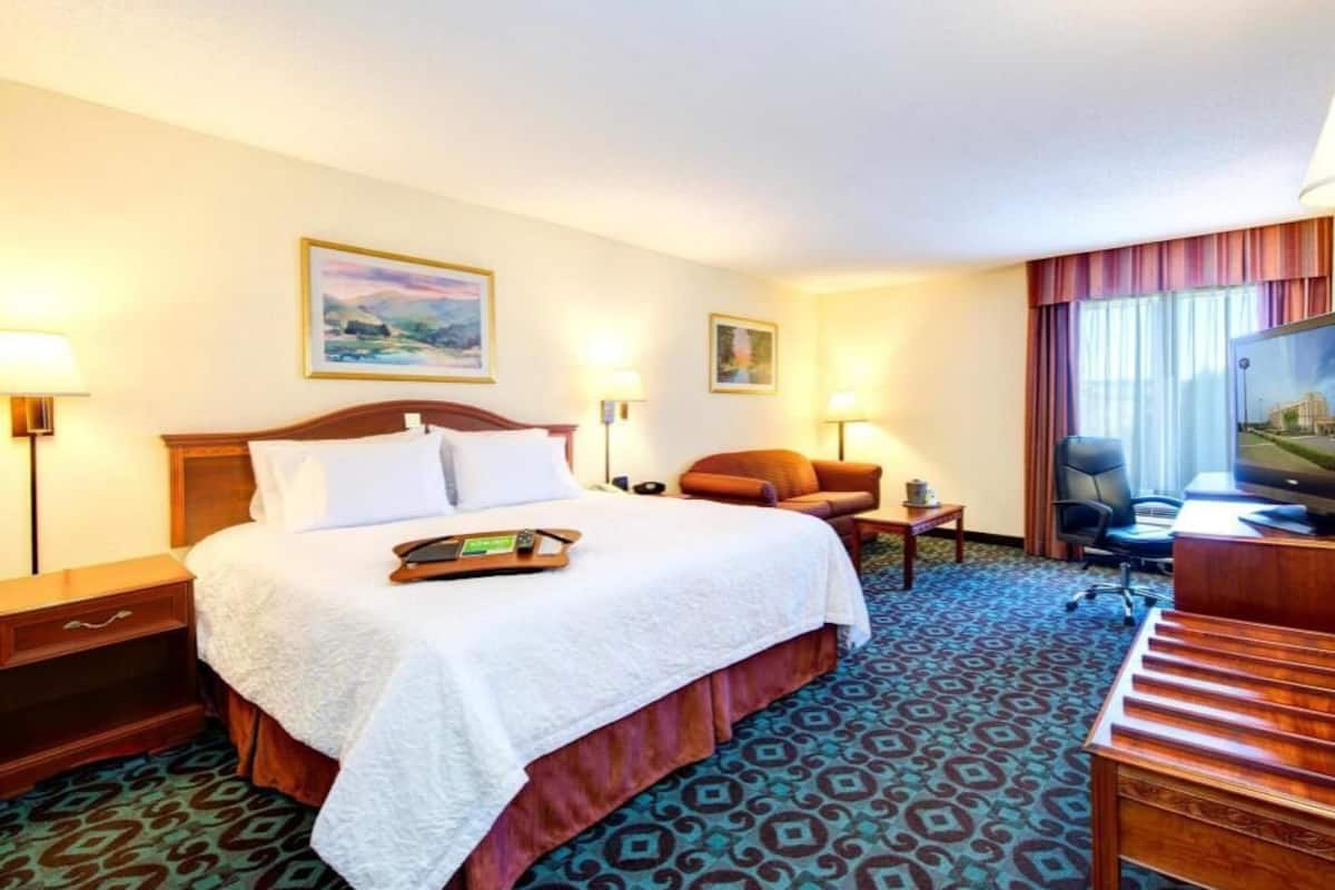 Habitación de hotel con cama, escritorio, silla, televisor y ventana con cortinas.