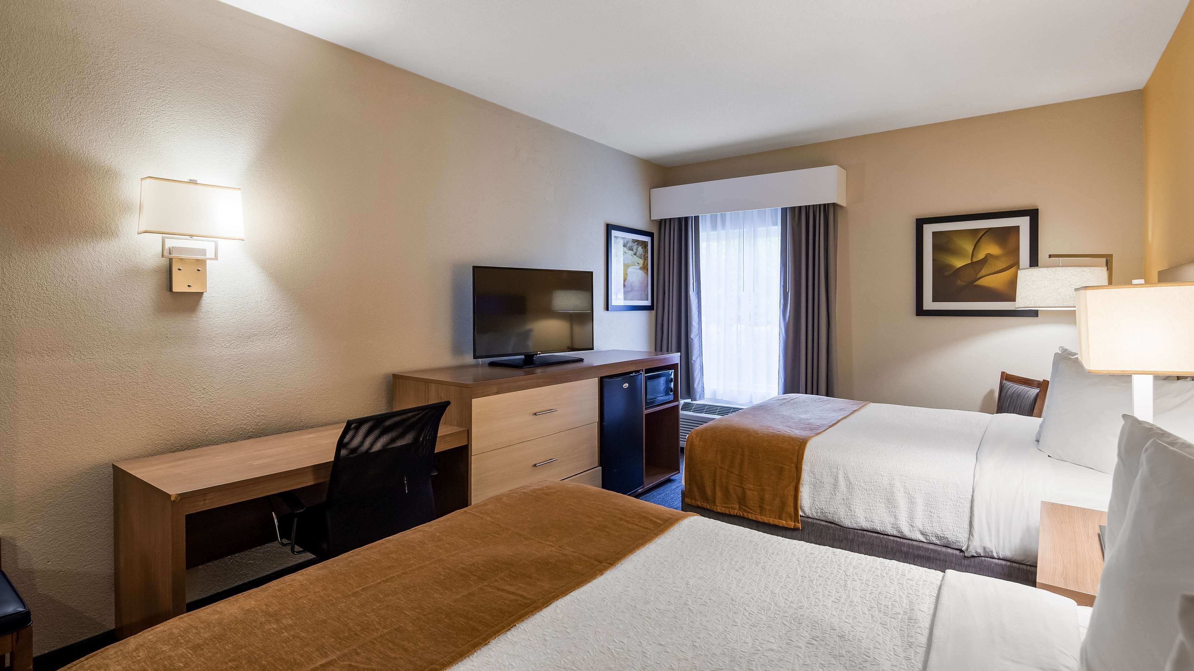Quarto de hotel com escrivaninha, duas camas, televisão e janela com cortinas.