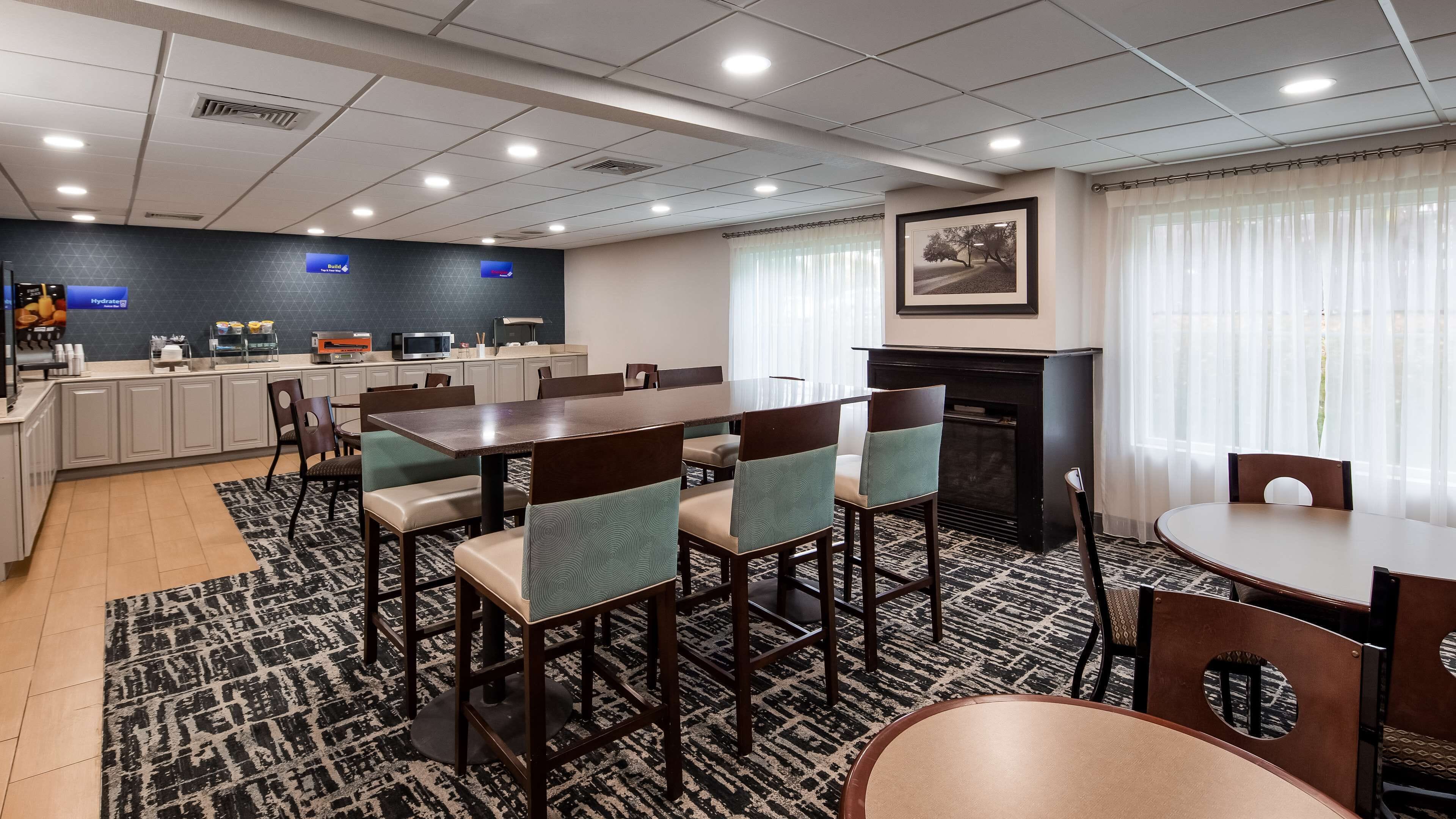 A hotel dining area with a long communal table, several chairs, a kitchen area with a microwave, and a fireplace.
