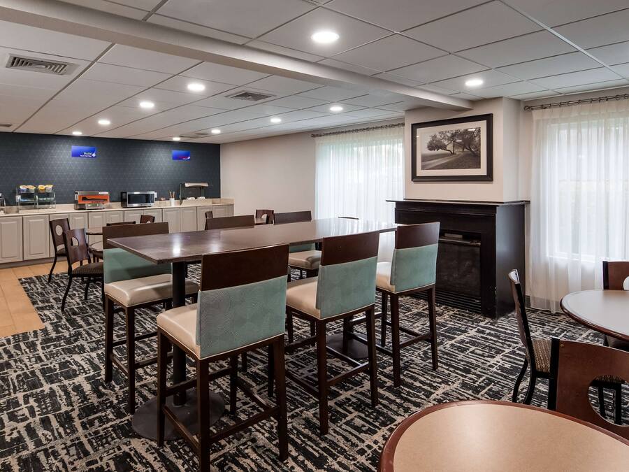 A hotel dining area with a long communal table, several chairs, a kitchen area with a microwave, and a fireplace.