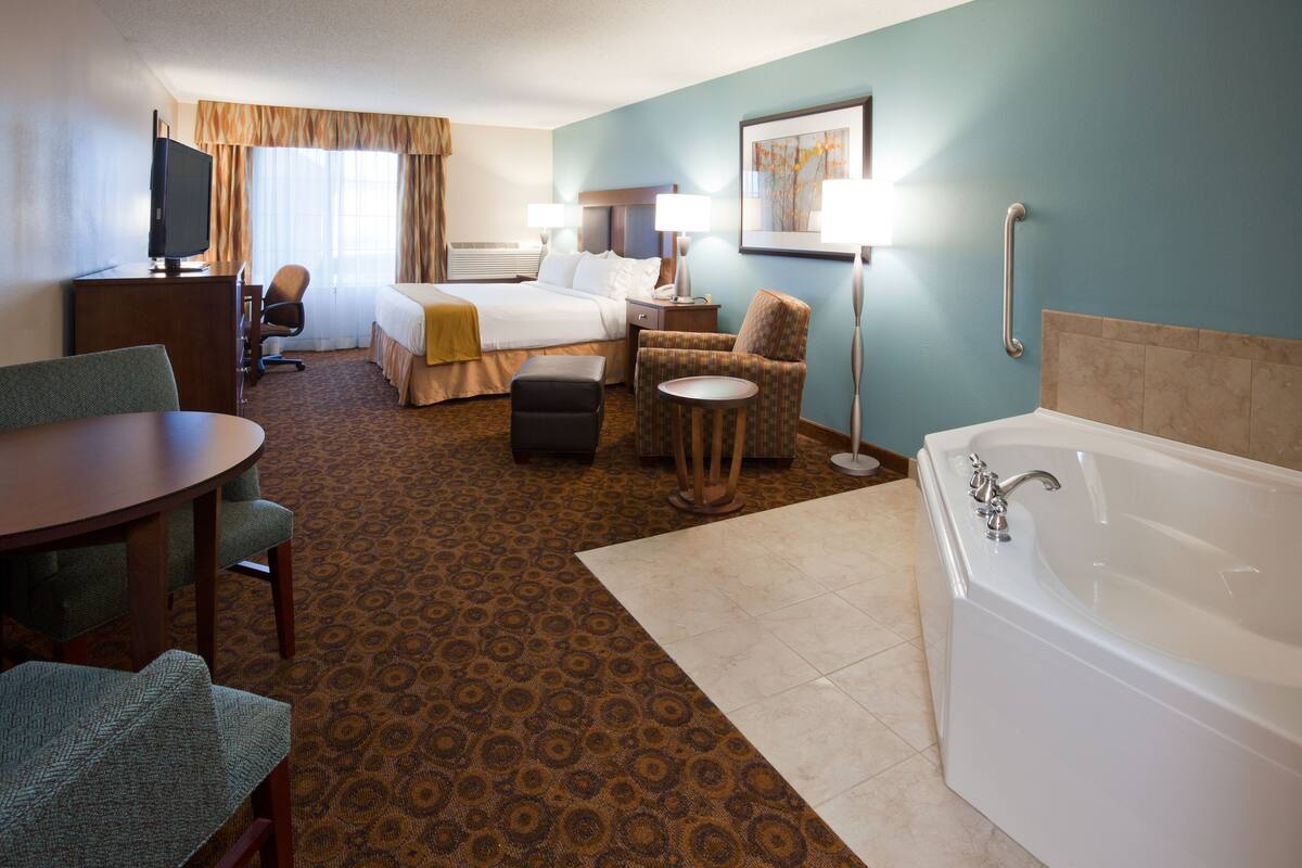 A hotel room with a bed, a desk with a television, a chair, a small table, a lamp, and a bathtub.