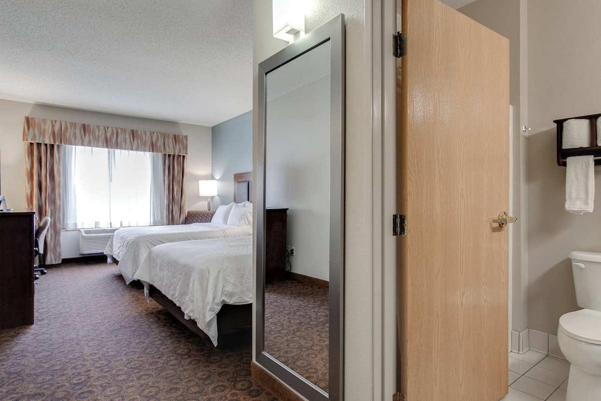 A hotel room with two beds, a bathroom with a toilet and towel rack, and a mirror reflecting the room.