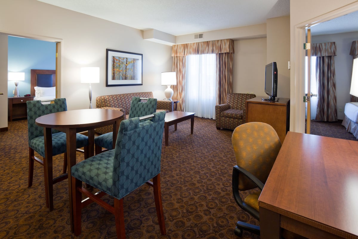 A hotel room with a dining table, four chairs, a sofa, a television, and a bed.
