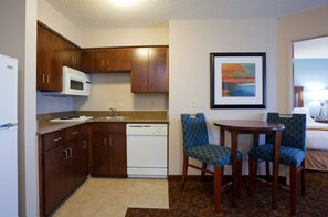 A compact kitchen with wooden cabinets, a white microwave, and a dishwasher. There is a framed painting on the wall and two blue upholstered chairs around a small table. - Holiday Inn Express Hotel & Suites St. Cloud by IHG (St Cloud)