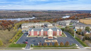 A multi-story hotel with a central courtyard, parking lot, and a view of a river and autumn foliage in the background. - Holiday Inn Express Hotel & Suites St. Cloud by IHG (St Cloud)